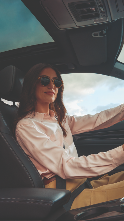 View from inside a car showing the driver's seat. A person wearing sunglasses and a light-colored top is seated behind the steering wheel. The car's interior features a sunroof and dark upholstery.